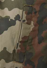 Camouflage fabric features shades of green, brown, and black with a textured surface. Visible stitching outlines a rectangular pocket detail.