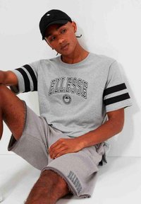 Camiseta gris con rayas negras en las mangas y logo "ELLESSE". Combinada con pantalones cortos grises con dobladillo deshilachado y marca lateral. Gorra negra.