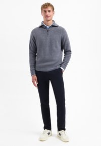 Man staat gekleed in een grijze rits-trui, lichtblauwe shirt, donkere broek en witte sneakers met marineblauwe details op een witte achtergrond.