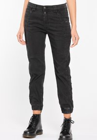 Černé zúžené džíny s hladkým povrchem, přední kapsy, zapínání na knoflíky a elastické manžety, doplněné černými šněrovacími botami.