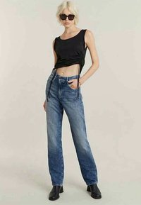 Žena sa kratkom plavom kosom koja nosi crni top bez rukava sa naborima, plave traperice viskog struka, crne čizme i tamne sunčane naočale stoji.