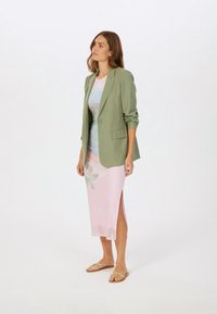Grüner taillierter Blazer über einem pastellfarbenen Tie-Dye-Kleid mit einem seitlichen Schlitz, kombiniert mit flachen Sandalen. Glatter Stoff und strukturiertes Design.