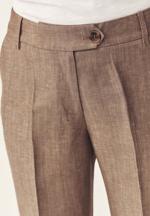 Nahaufnahme von braunen Leinenhosen mit Knopfverschluss und Bundfalten, getragen zu einem weißen Oberteil.