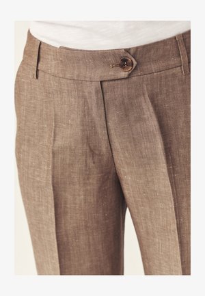 Nahaufnahme von braunen Leinenhosen mit Knopfverschluss und Bundfalten, getragen zu einem weißen Oberteil.