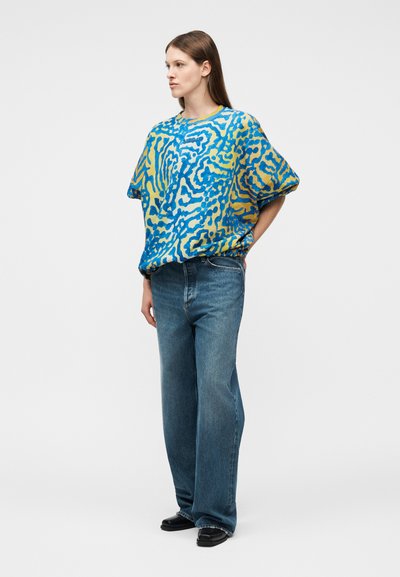 Γυναίκα με μακριά καστανά μαλλιά, φορώντας άνετο πολύχρωμο πουλόβερ με μπλε και κίτρινο μοτίβο, φαρδιά μπλε τζιν παντελόνια και μαύρα παπούτσια, στέκεται με τα χέρια πίσω.