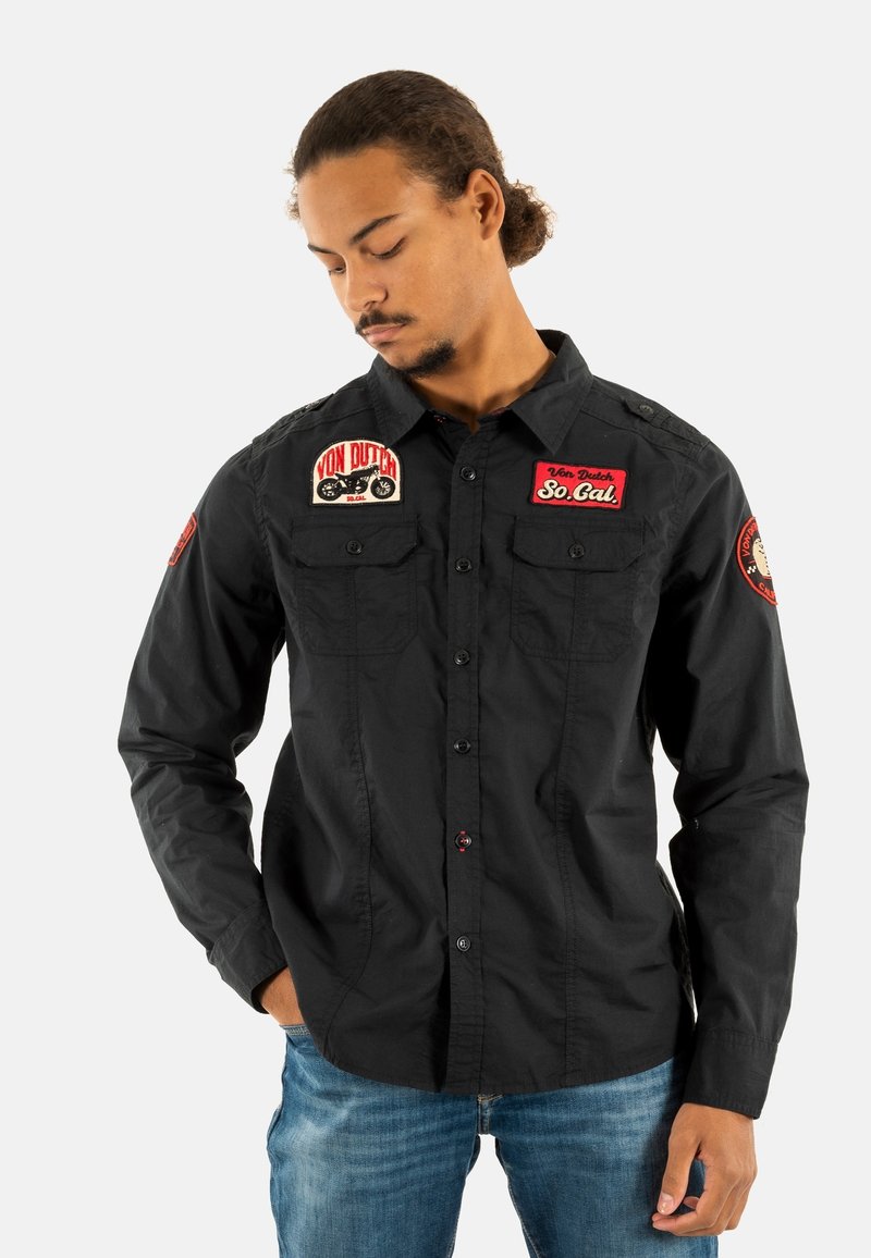 Camisa negra de manga larga con parches. Cuenta con cuello tipo botón, dos bolsillos en el pecho y acentos bordados en rojo. Tela suave y ligera.