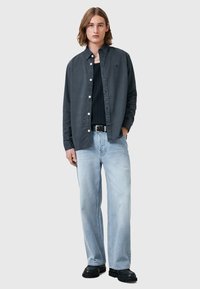 Camicia grigia a maniche lunghe con bottoni sopra una maglietta nera a costine, abbinata a jeans azzurri morbidi e scarpe nere chunky.
