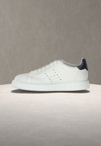 Baskets basses blanches avec motif latéral perforé, semelle épaisse rainurée, lacets blancs et accent noir au talon sur fond neutre.