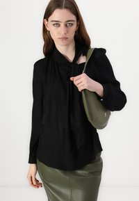 Svart bluse med sløyfedetalj, lange ermer og subtil tekstur. Kombinert med et figurnært olivengrønt skinnskjørt og en liten grønn veske.