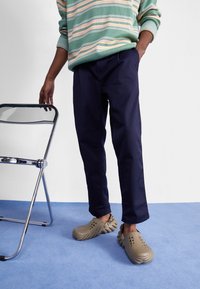 Zelený svetr s pruhy, tmavě modré kalhoty a béžové texturované dřeváky s ventilačními otvory. Vedle na modrém povrchu je chromová skládací židle.