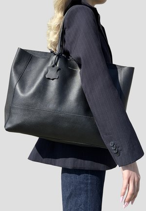 Donna con capelli biondi lunghi, indossa un blazer scuro a righe sottili e jeans, porta una grande borsa tote nera in pelle texturizzata sulla spalla.