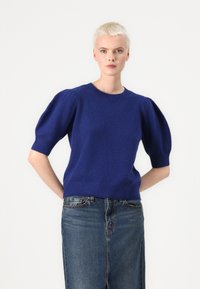 Pull en maille bleu royal avec des manches bouffantes courtes et un col rond, associé à une jupe en jean, sur un fond blanc uni.
