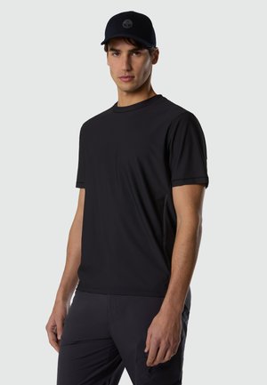 Hombre con una camiseta negra de manga corta lisa, pantalones oscuros y una gorra de béisbol negra con un logo circular, posando frente a un fondo gris claro.