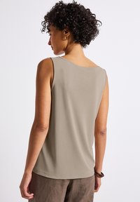 Ärmlös topp i mjukt, lätt taupe-tyg med rundad halsringning och en något böjd nederkant, kombinerad med lösa bruna byxor.