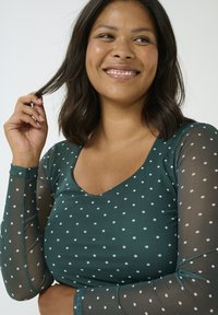 Teal-farbene Bluse mit Polka-Dots aus leichtem Stoff und langen, durchsichtigen Ärmeln. Mit einem V-Ausschnitt und kleinen weißen Punkten versehen. Sanfte Textur.