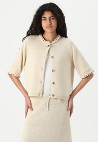 Cardigan crème en tricot à manches courtes, avec un col tressé et des boutons en bois. Tissu texturé avec une coupe ample, associé à une jupe assortie.
