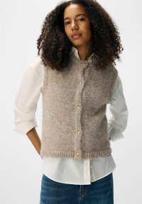 Strickweste aus weichem beigen Garn, mit rundem Halsausschnitt, sechs goldenen Knöpfen und einem gerippten Saum, getragen über einem weißen Hemd mit Kragen.