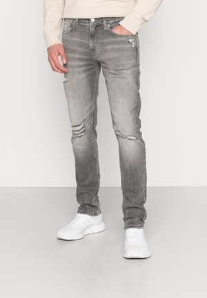 Mand iført falmede grå revne jeans og hvide sneakers med den ene hånd i lommen mod en ensfarvet baggrund.
