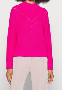 Pull en tricot fuchsia avec un col haut, doté d'un motif texturé et d'accents de points ouverts, associé à un pantalon de couleur claire.