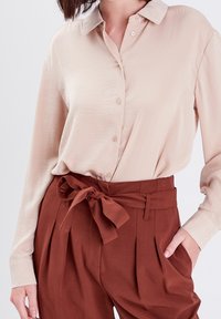 Chemise beige boutonnée en tissu lisse avec un col classique, associée à un pantalon marron taille haute avec une ceinture nouée et des plis.