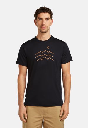 Mężczyzna w czarnej koszulce z brązowym nadrukiem góry i słońca, ubrany w beżowe spodnie, stoi na jasnym, jednolitym tle.