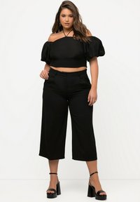 Sort off-shoulder crop top med puffærmer, parret med højtaljede, brede sorte bukser og kraftige sorte platformhæle.