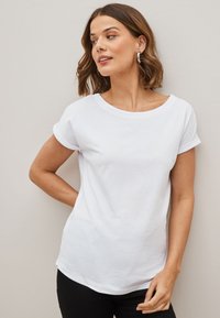 Femme aux cheveux courts et ondulés, portant un t-shirt blanc à manches courtes et un pantalon noir, regardant sur le côté sur un fond uni.