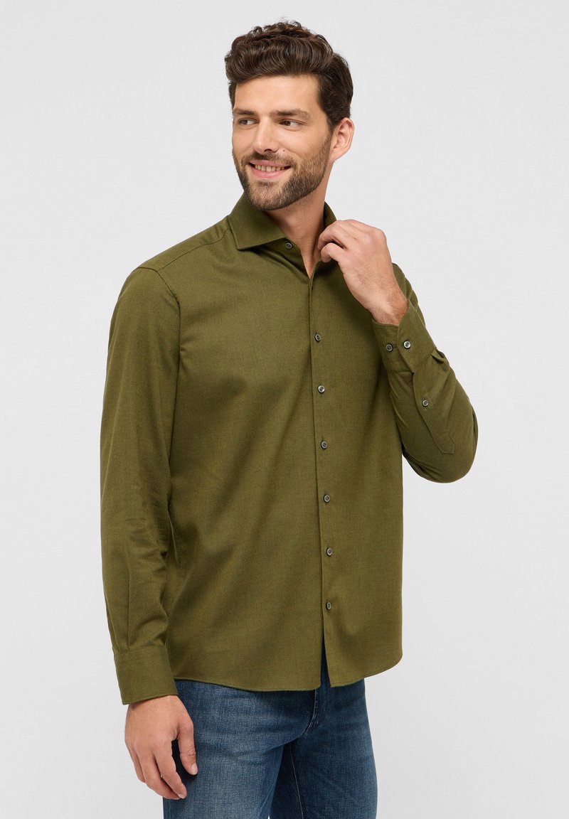 Camicia a maniche lunghe verde oliva con colletto classico, chiusura frontale con bottoni e polsini con bottoni. Realizzata in tessuto texturizzato, abbinata a jeans blu.