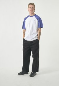 White and purple T-shirt with short raglan sleeves, black pants, and black sneakers. Features a small logo on the chest and loose fit.