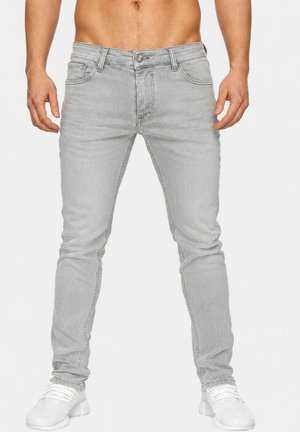 Man draagt lichtgrijze slim-fit jeans en witte sneakers, staat met ontspannen handen langs zijn zij voor een effen achtergrond.