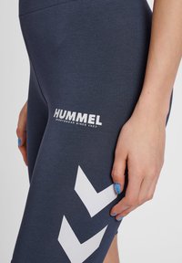 Marinblå leggings i stretchmaterial med en vit "HUMMEL"-logotype och ett vitt chevronmönster på sidan. Slät textur, åtsittande design.