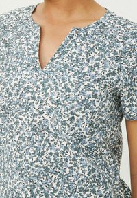 Kurzarm-Top mit Blumendesign und V-Ausschnitt. Der Stoff zeigt Blau- und Grüntöne auf einem cremefarbenen Hintergrund mit feinen Details.