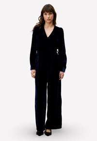 Combinaison en velours bleu marine avec un col en V, des manches longues et une taille nouée. Design à jambes larges avec une texture douce et un éclat subtil.