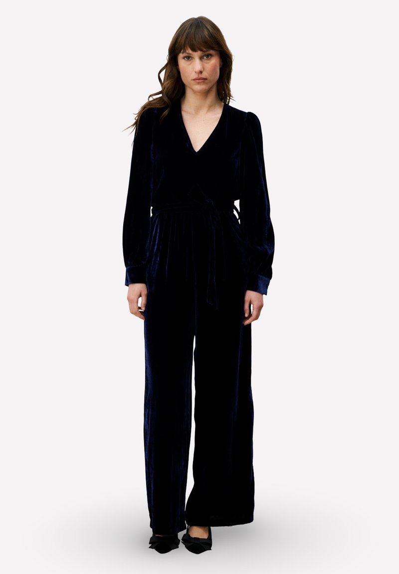 Combinaison en velours bleu marine avec un col en V, des manches longues et une taille nouée. Design à jambes larges avec une texture douce et un éclat subtil.