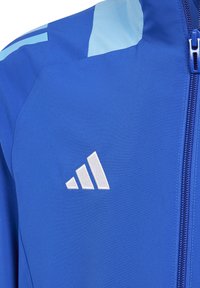 Königblaue Jacke mit hellblauen Akzenten, ausgestattet mit einem Reißverschluss und einem gestickten weißen Markenlogo auf der Brust. Glattes, leichtes Material.