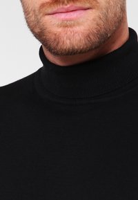 Gros plan du bas du visage et du cou d’un homme portant un pull à col roulé noir, montrant une barbe rousse taillée et des lèvres.