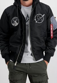 Musta bomber-takki harmailla yksityiskohdilla, jossa on Apollo 11- ja planeettakirjailu, vetoketjusuljenta ja kangasmerkki hihassa.