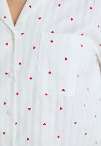 Camicia bianca con motivo a righe sottili e piccoli cuori rossi, dotata di taschino sul petto, realizzata in tessuto morbido e leggero.