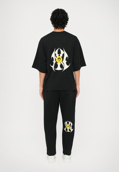 Sort oversized t-shirt og sæt af sweats, der har hvide flammeagtige mønstre samt et gult smilende ansigt på ryggen og venstre ben.