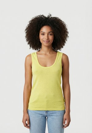 Giovane donna con capelli ricci che indossa una canottiera gialla e jeans azzurri, in piedi davanti a uno sfondo bianco semplice.