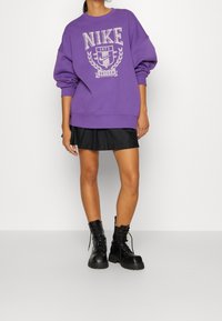 Lila Nike-tröja med grafiskt tryck, avslappnad passform, ribbade ärmslut, matchad med en svart plisserad kjol och svarta kängor.