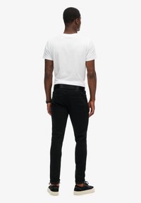 Jeans negros ajustados con una textura suave, combinados con una camiseta blanca lisa y zapatillas negras informales. Vista desde la parte trasera.