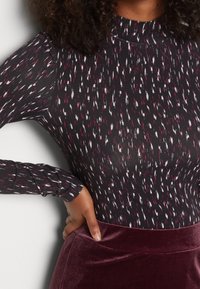 Langärmliger Bodysuit in Schwarz mit verstreutem roten und weißen Muster; weicher, elastischer Stoff; Rundhalsausschnitt; kombiniert mit einem burgunderfarbenen Samtrock.