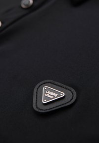 Sort polo skjorte med en triangulær læderpatch med prægede bogstaver "OMBRA EXIMO" og dekorativ syning nær kraven.