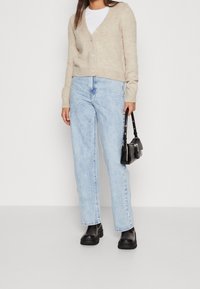 Woman wearing beige cardigan, white top, light blue jeans, black boots, holding a small black handbag, standing against a plain background.