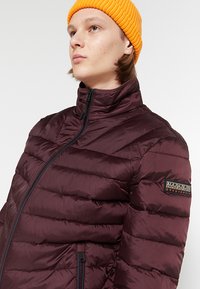 Giacca imbottita bordeaux con design a cuciture orizzontali, colletto alto e chiusura con zip. Presenta un patch con logo sulla manica e una texture liscia.