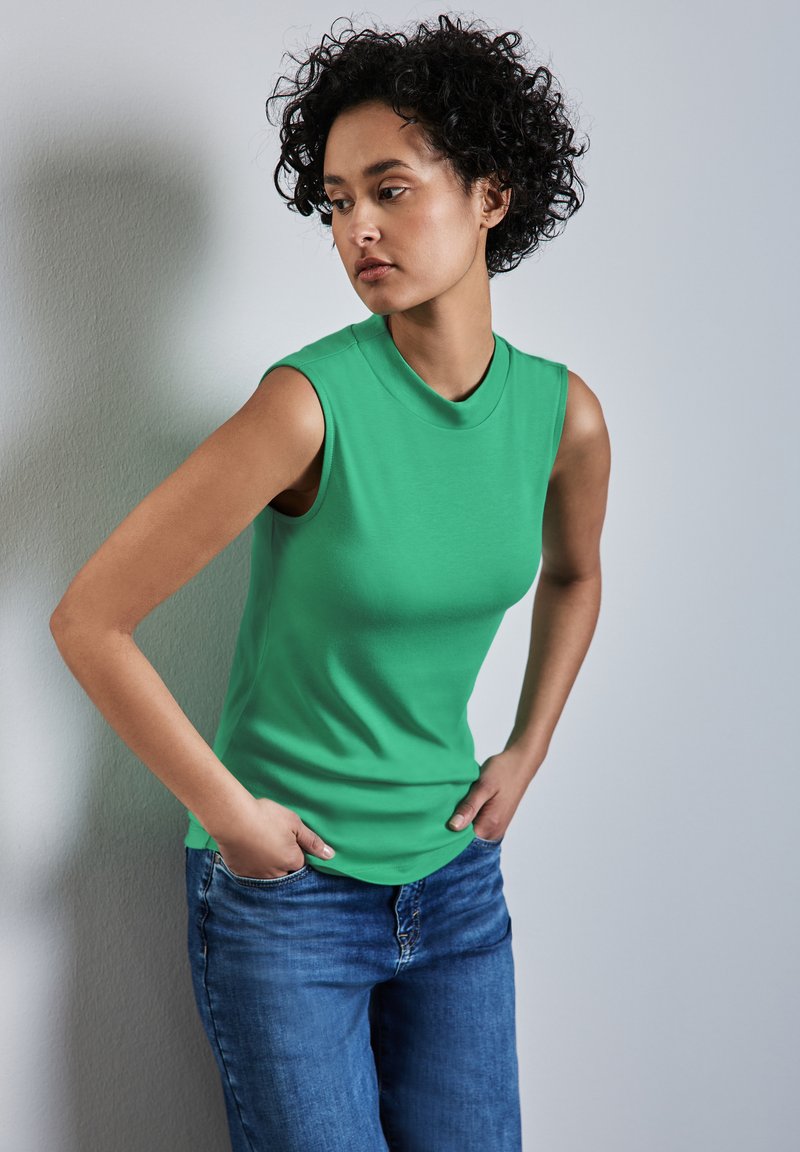 Top verde senza maniche realizzato in tessuto morbido, con un collo alto. Abbinato a jeans in denim blu. Design semplice senza motivi o dettagli.