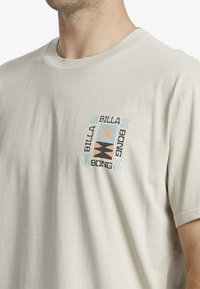 Camiseta de algodón beige claro con un estampado gráfico multicolor que presenta el texto "BILLA BONG" y formas geométricas centradas en el pecho.