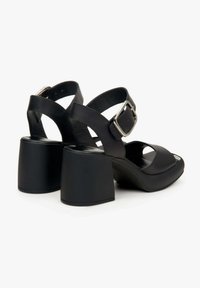 Zwarte gehakte sandalen met een brede blokhak, gespsluiting, open teen en kruislings straps ontwerp; gemaakt van glad kunstleer.