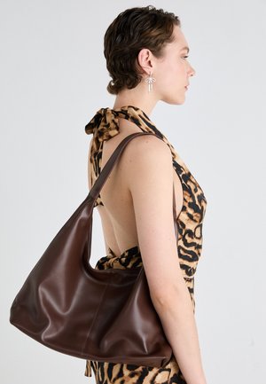 Žena s krátkými vlasy, oblečená v leopardím vzorovaném šatech s halter výstřihem a stříbrnými náušnicemi ve tvaru mašle, nese velkou hnědou koženou kabelku přes rameno.